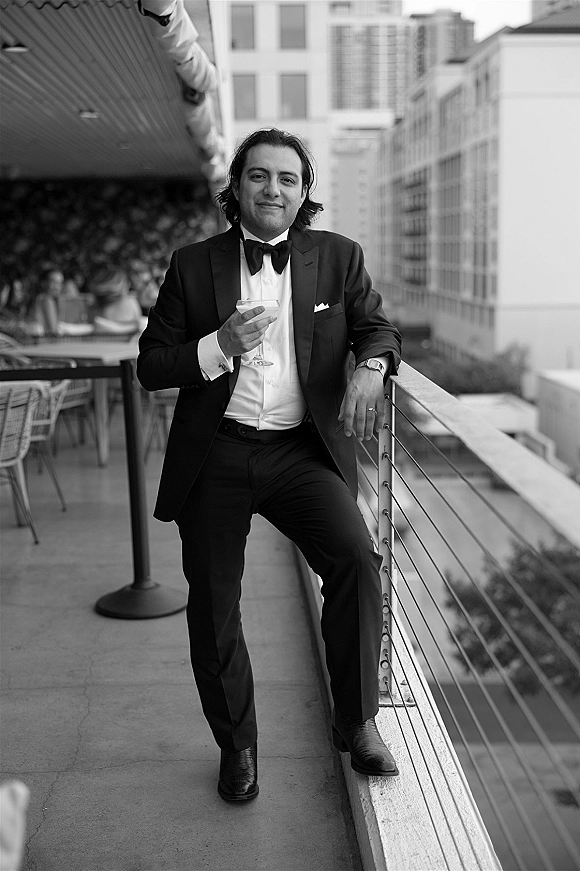 Groom portrait in a black tie tuxedo, leaning on a rooftop terrace railing with a cocktail glass, city skyline behind him
