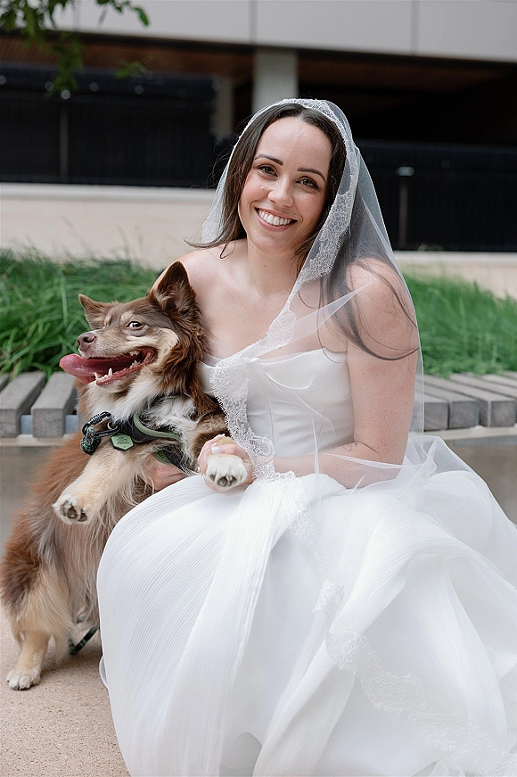 Bridal portrait of a bride with dog, wearing a lace-trim veil and strapless gown, sitting on outdoor concrete steps by greenery