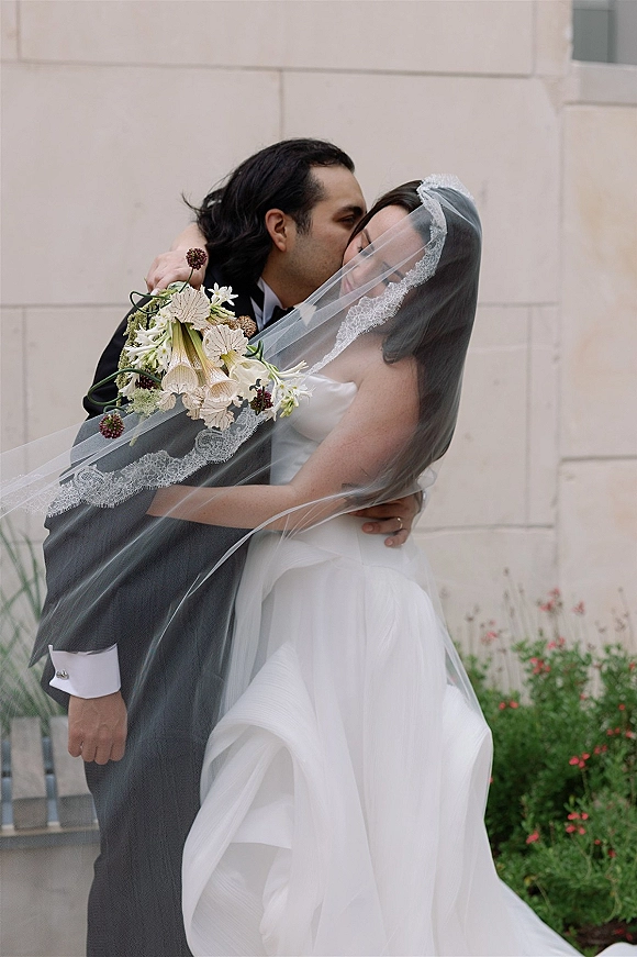 Wedding kiss portrait of the bride and groom kissing under a lace veil, bouquet in hand, against a stone wall with garden greenery