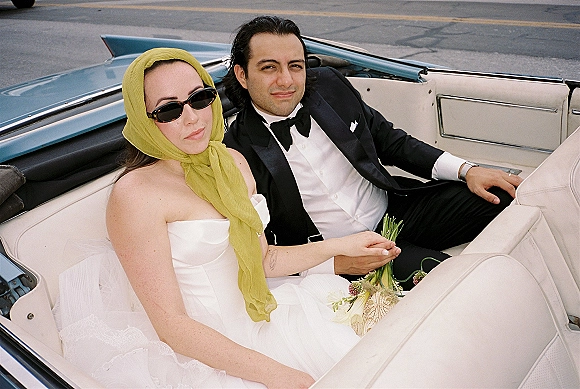 Couple portrait of bride and groom in car, riding a vintage convertible with bouquet, headscarf, and sunglasses on city street