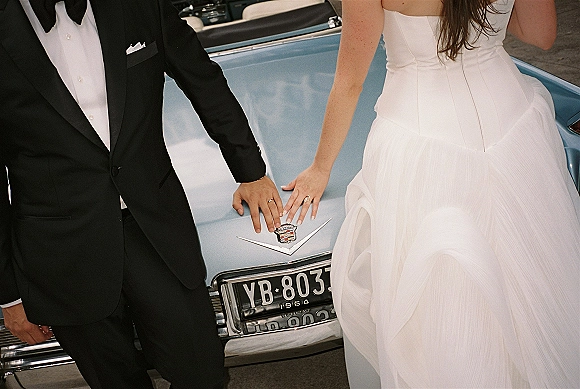 Couple portrait with bride and groom hands resting on a vintage car hood ornament, wedding rings visible against street pavement