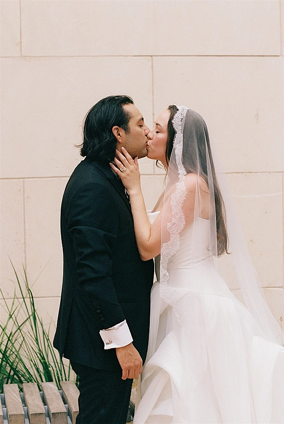 Wedding kiss as bride and groom kiss in side profile, bride touching his face with lace veil and ring against white stone wall greenery