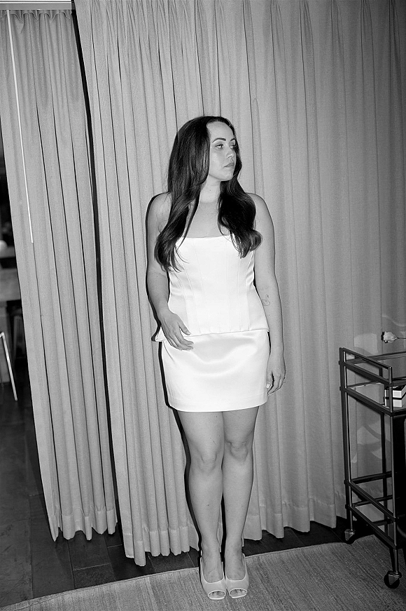 Bridal portrait in black and white of a bride in a short wedding dress with strapless corset bodice, posing by a curtain backdrop and bar cart