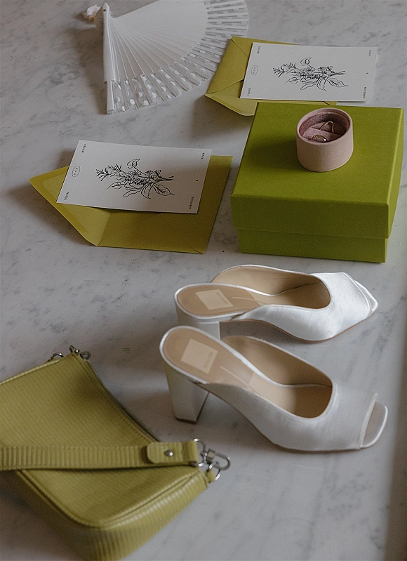 Wedding details flatlay with a wedding invitation suite, rings in a box, white heeled mules, green clutch, and fan on marble background