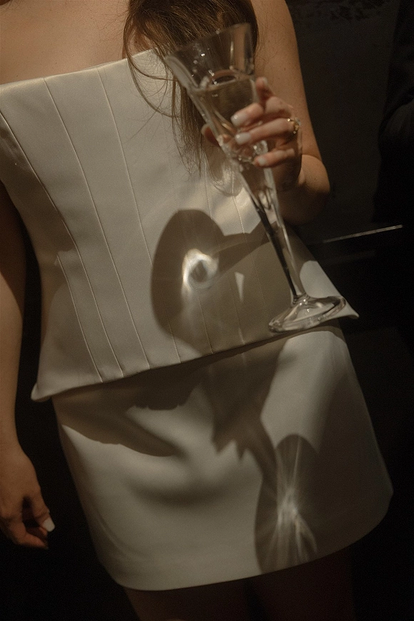 Wedding guest dress in a white strapless mini dress, holding a champagne flute with ring and manicured nails against a dark interior background