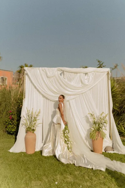 Bridal portrait of a bride holding a long stem white bouquet in a strapless satin gown, before a white draped backdrop outdoors