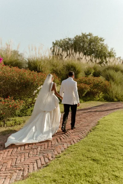 Couple portrait of bride and groom walking away hand in hand, her veil and long train trailing on a brick garden path