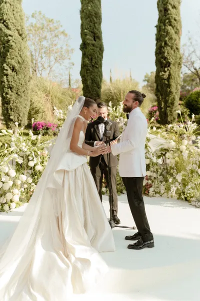 Wedding vows as bride and groom hold hands, her veil flowing beside a microphone stand amid white garden florals under blue sky