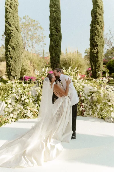 Wedding kiss portrait of bride and groom kissing, her long veil flowing beside a floral-lined aisle in an outdoor garden with cypress trees