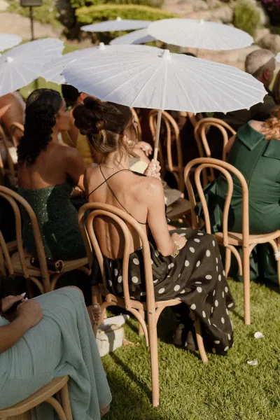 Wedding guests seated on wooden bentwood chairs, holding white paper parasols on a sunny lawn near stone steps, seen from behind
