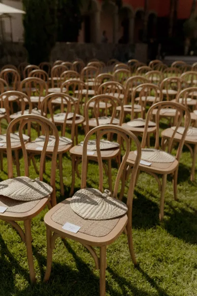 Ceremony seating with outdoor wedding chairs in neat rows, wood chairs topped with straw hats and place cards in a grassy courtyard