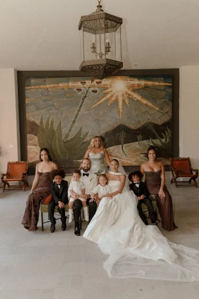 Wedding family portrait of bride and groom with kids on a sofa, veil draped over train, under chandelier before a mural wall in lobby