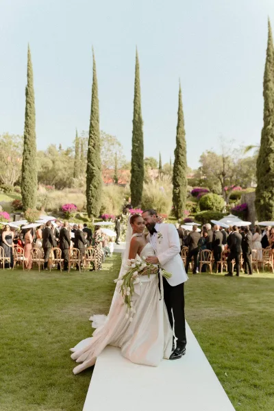 Couple portrait of bride and groom embracing, bride in strapless dress with veil holding bouquet on a garden aisle with cypress trees