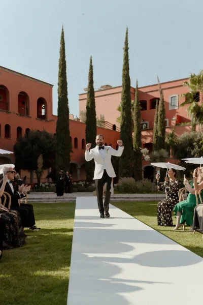 Wedding recessional as groom in a white tuxedo jacket walks the aisle runner, guests cheering under parasols in a sunny courtyard with cypress trees