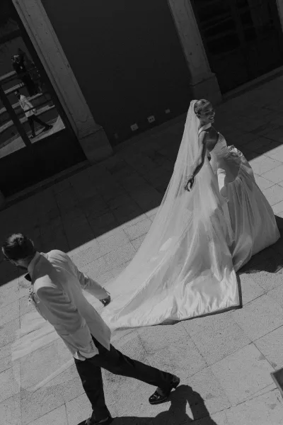 Couple portrait of bride and groom walking in a stone courtyard, bride looking back while holding her dress train in dramatic shadows