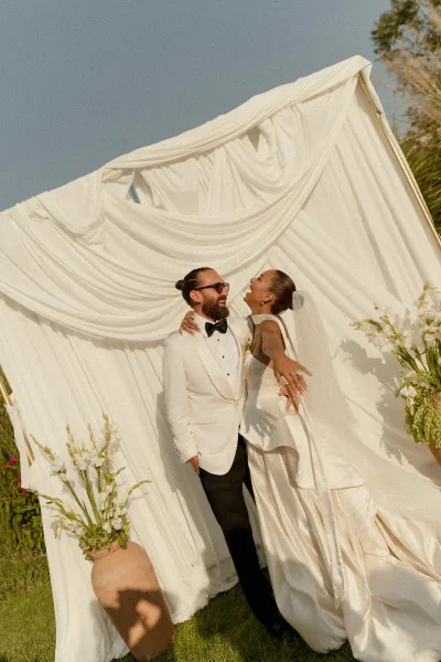 Couple portrait of bride and groom laughing, her strapless dress and his white tuxedo jacket with sunglasses on a lawn by trees