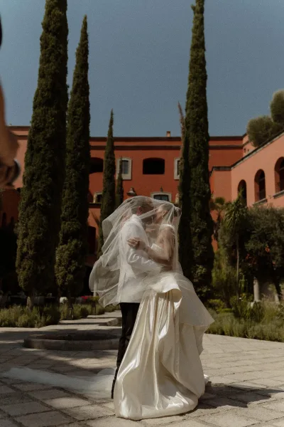 Wedding kiss portrait of bride and groom under a veil, embracing in a sunlit courtyard with cypress trees and terracotta building backdrop