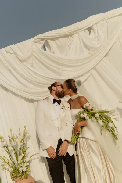 Wedding kiss portrait of bride and groom kissing, her veil and strapless gown framed by a white tulip bouquet and open sky backdrop