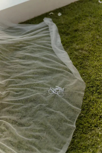 Wedding veil with cathedral wedding veil length spread on a grass lawn, sheer tulle showing an embroidered monogram near a white structure