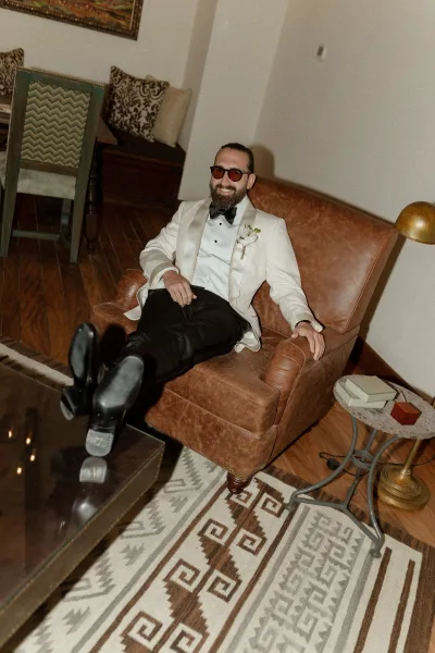 Groom portrait in a white tuxedo lounging in a leather armchair, wearing sunglasses and boutonniere in a modern indoor lounge setting