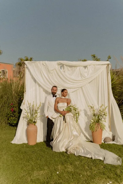 Couple portrait of bride in a strapless satin gown holding a bouquet beside groom in white tuxedo, before draped backdrop on lawn with palms