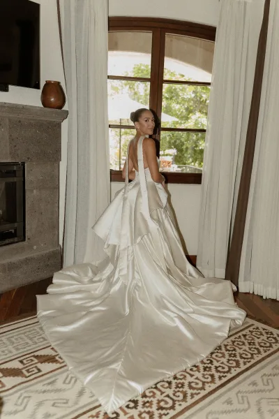 Bridal portrait of a bride in a satin wedding dress with a long train, looking over her shoulder by an arched window with curtains
