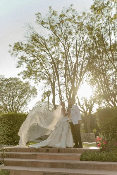 Wedding couple portrait of bride and groom holding hands on sunlit stone steps in a garden, cathedral veil blowing as she holds a bouquet