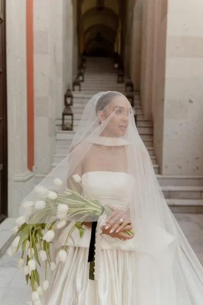Bridal portrait of a bride with veil holding a white tulip bouquet on a stone staircase with lanterns and an archway behind her