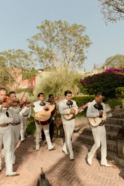 Mariachi band in white charro suits walks down a brick garden walkway with violins and guitars under a bright blue sky