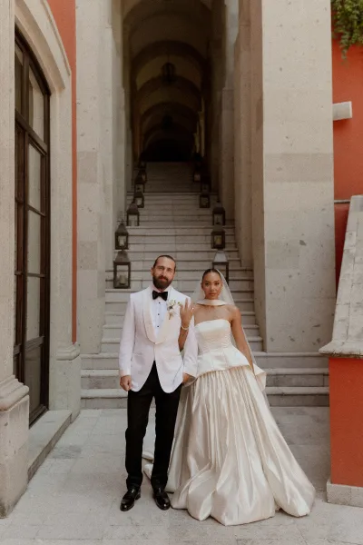Couple portrait of bride in strapless gown and veil with groom in white dinner jacket on a stone staircase in an arched corridor with lanterns