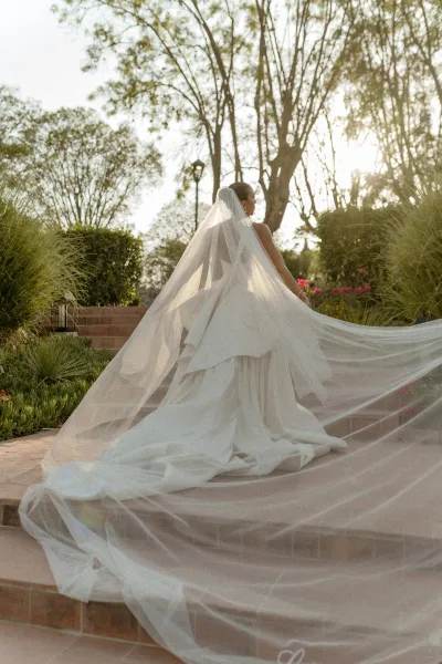 Bridal portrait of a bride in a wedding dress with long train, cathedral veil flowing behind her as she walks up sunlit garden stone steps