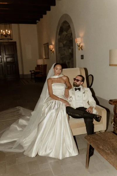 Couple portrait of bride in strapless satin ballgown and veil leaning on groom in white tuxedo with sunglasses in arched hallway