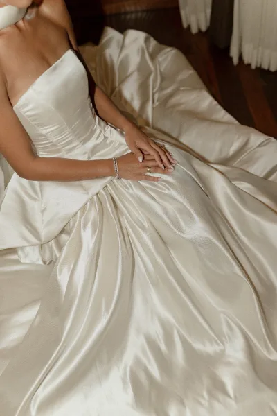 Wedding dress close-up shows a bride’s hand on a strapless satin gown, diamond bracelet and rings visible on a wood floor near hanging dresses