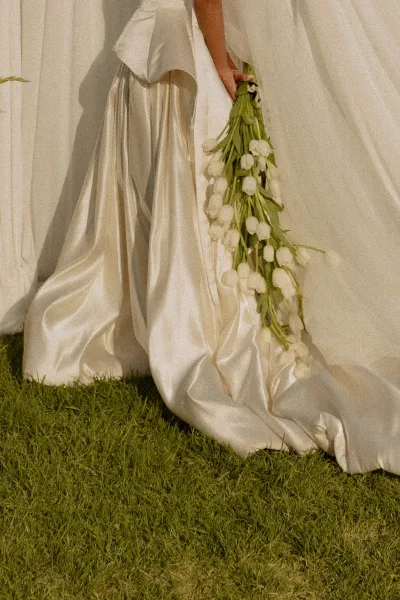 Bridal bouquet of white tulips with long stems held against a satin wedding dress and trailing train on a grass lawn by white drapes
