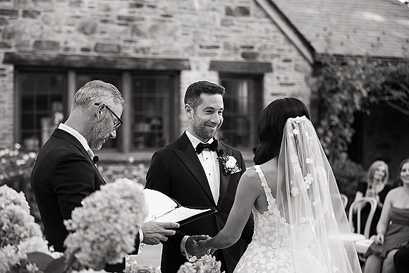 Wedding vows as bride in lace dress and veil holds hands with groom in tux, officiant reading beside microphones in brick courtyard