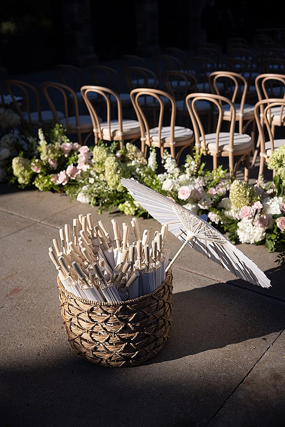 Ceremony aisle decor with ground floral aisle runner lining a stone patio, with wooden chairs, wicker fan basket, and white parasol accent