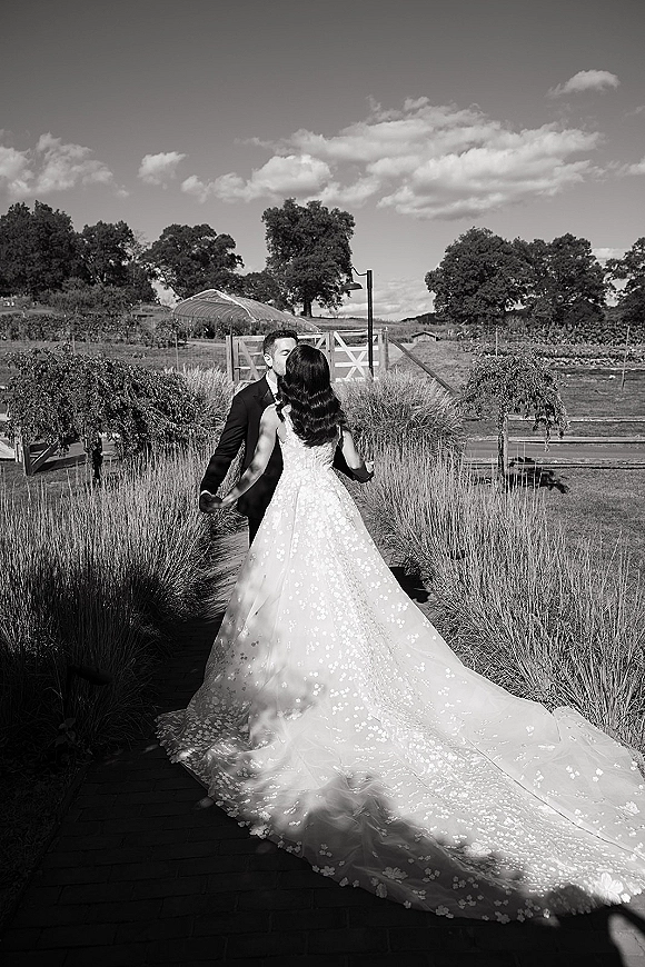Wedding kiss portrait of bride and groom kissing, holding hands on a wooden walkway, her lace train flowing beside vineyard rows under clouds