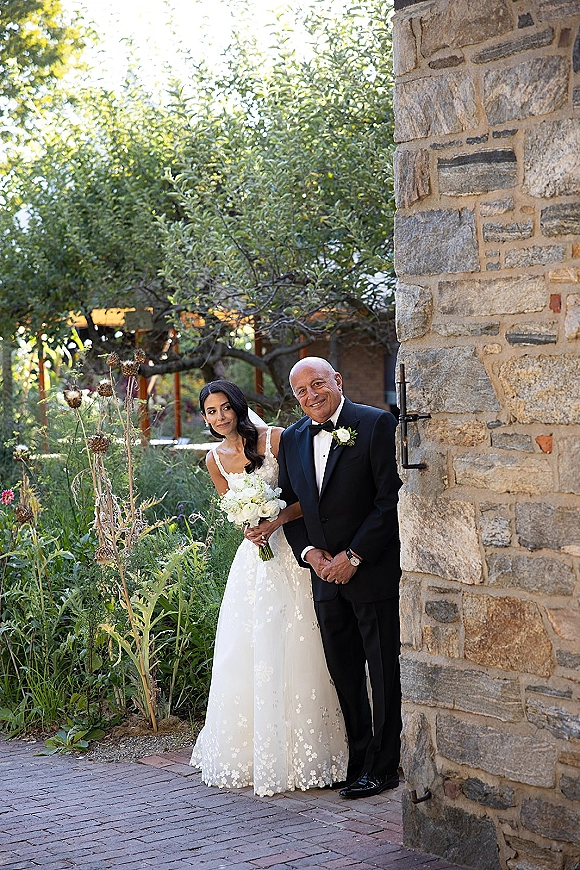 Couple portrait of bride and groom peeking, bride holding a white bouquet beside a stone wall with garden greenery and pergola behind