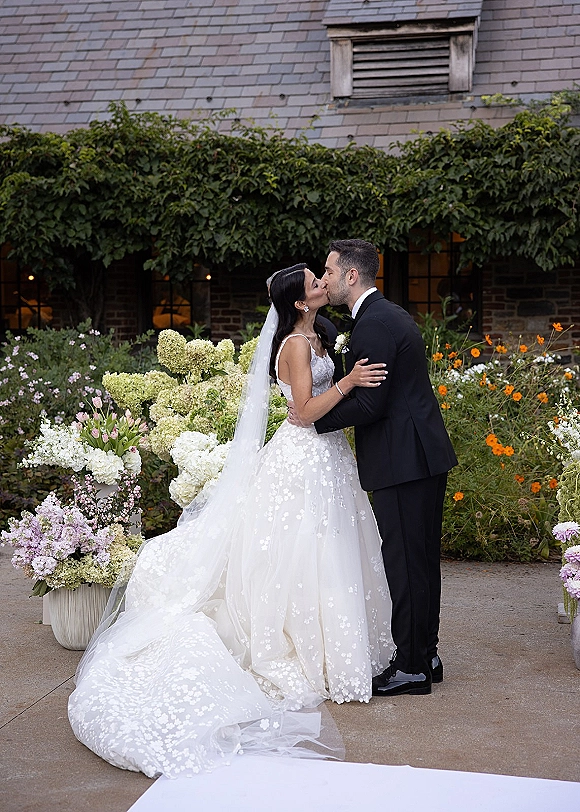 Wedding kiss portrait of bride and groom kiss, her long veil trailing by oversized florals on a garden patio with ivy-covered building backdrop