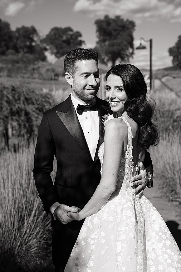 Couple portrait in a black and white wedding portrait style, bride in lace strap gown and groom in tuxedo embracing in a field under clouds