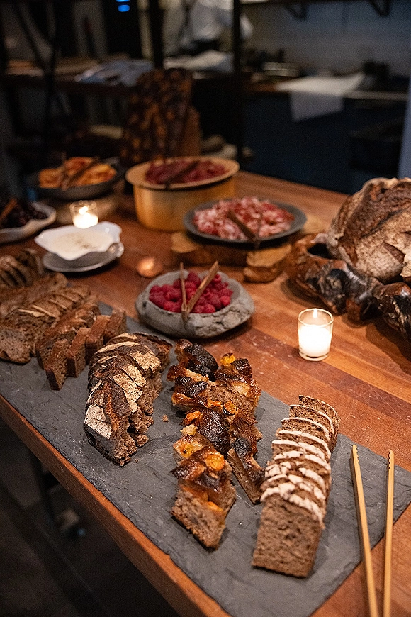 Wedding dessert table with loaf cake, sliced bread, raspberries, and crackers on a wooden table with votive candles in a dim reception space