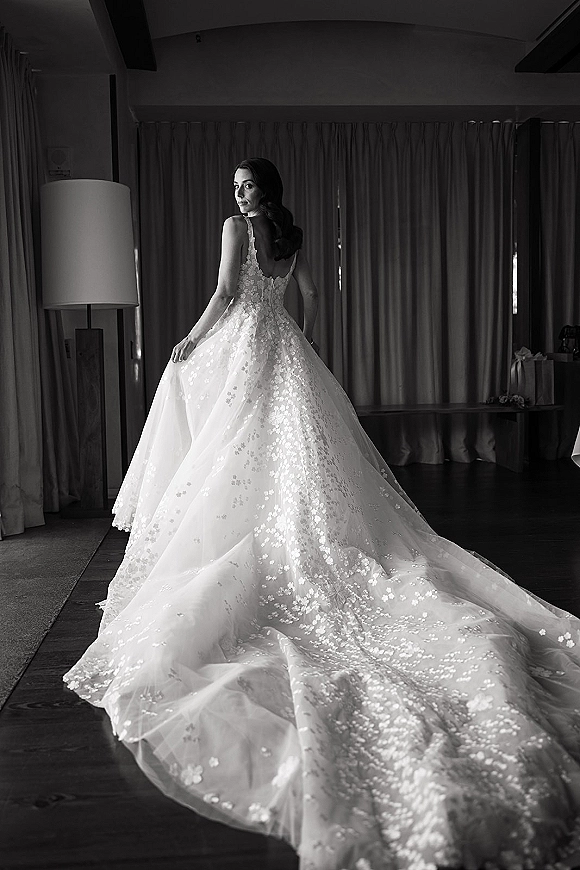 Bridal portrait of a bride in ball gown, showing a backless lace dress with tulle skirt and train in a hotel room by curtains and lamp