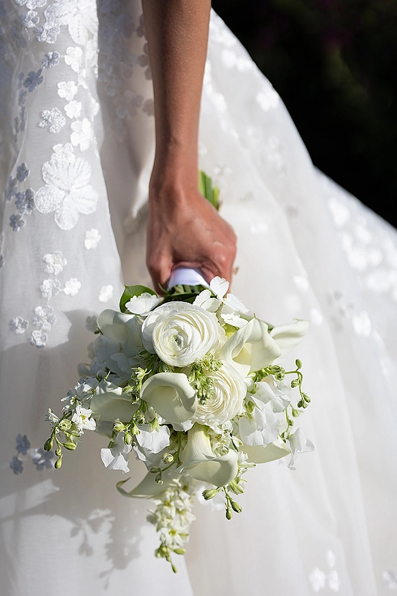 Bridal bouquet of white calla lilies and ranunculus wrapped with a white ribbon, held against a lace-appliqué gown in sunlit greenery