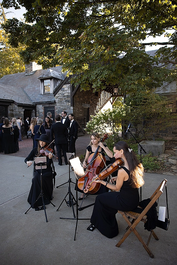 Wedding cocktail hour with a wedding string trio in black dresses playing violins and cello in a brick courtyard by a stone building