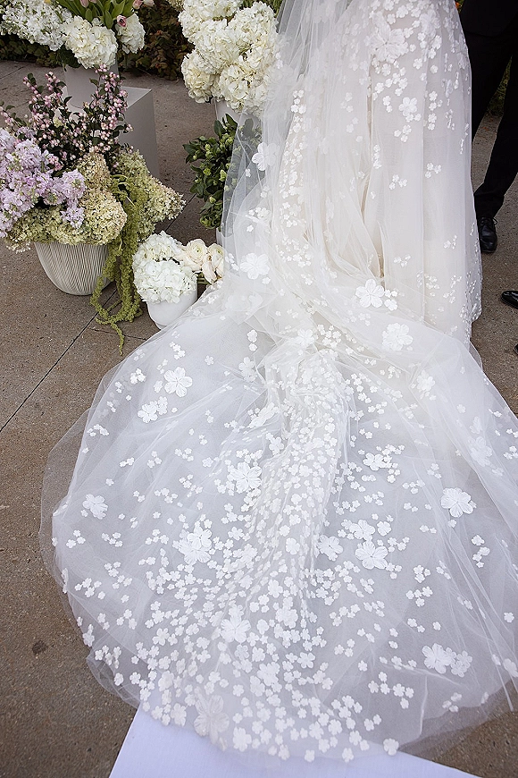 Wedding dress train with floral appliqué veil trailing over a tulle skirt beside hydrangea and rose arrangements on stone pavement
