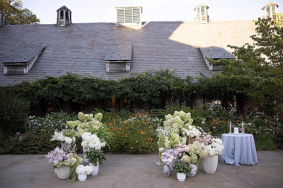 Ceremony altar decor with lush hydrangea wedding floral altar in urns and vases, plus pillar candles on a stone patio by an ivy brick wall