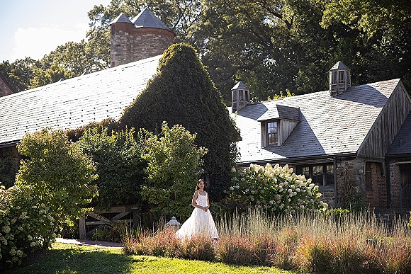 Bridal portrait of a bride in a lace bodice wedding dress with tulle skirt, standing by an ivy-covered stone building and hydrangeas
