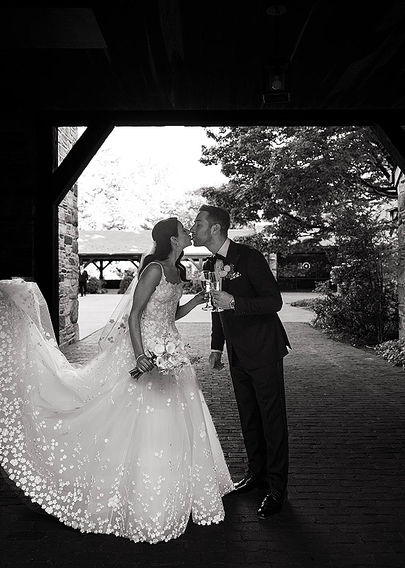 Wedding kiss portrait of bride and groom kissing, holding champagne flutes beneath a stone archway in a brick courtyard with trees