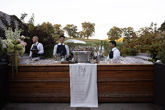 Wedding bar setup with a wood bar, cocktail menu sign, liquor bottles, and champagne flutes in an open field with trees behind