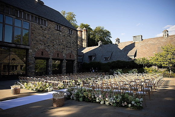 Ceremony setup with outdoor wedding ceremony seating, wood chairs flanking a white aisle runner and lush florals in a stone courtyard patio